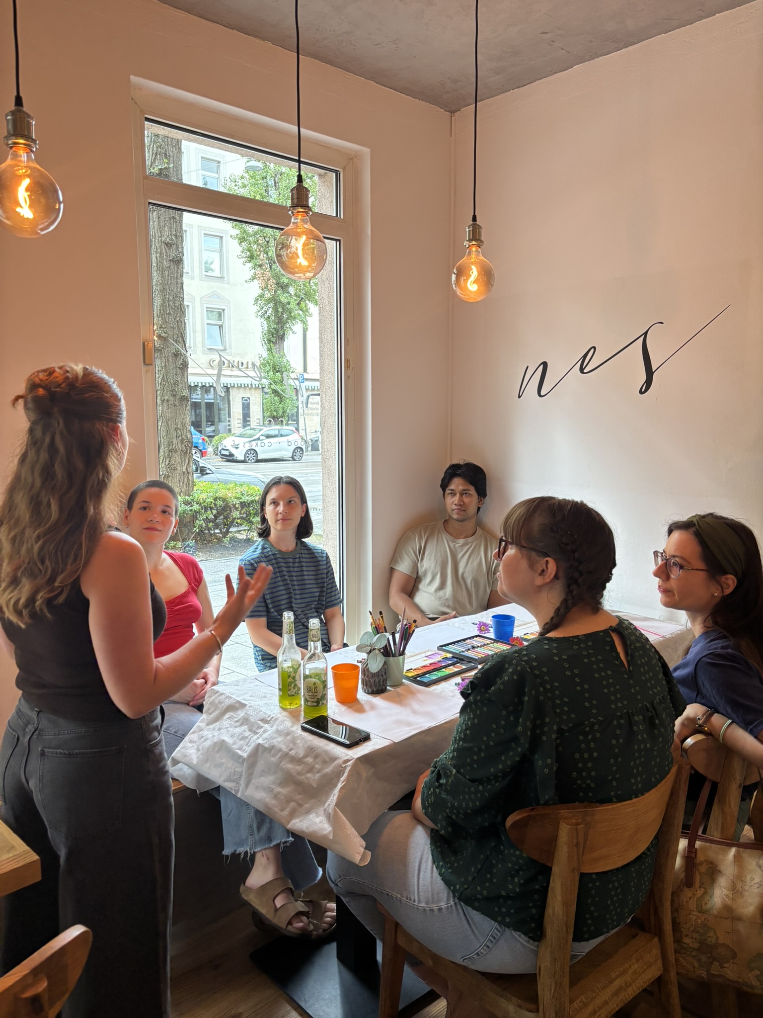 Workshop leader standing and talking to participants seated around a cafe table covered with watercolors, pastels and colored pencils